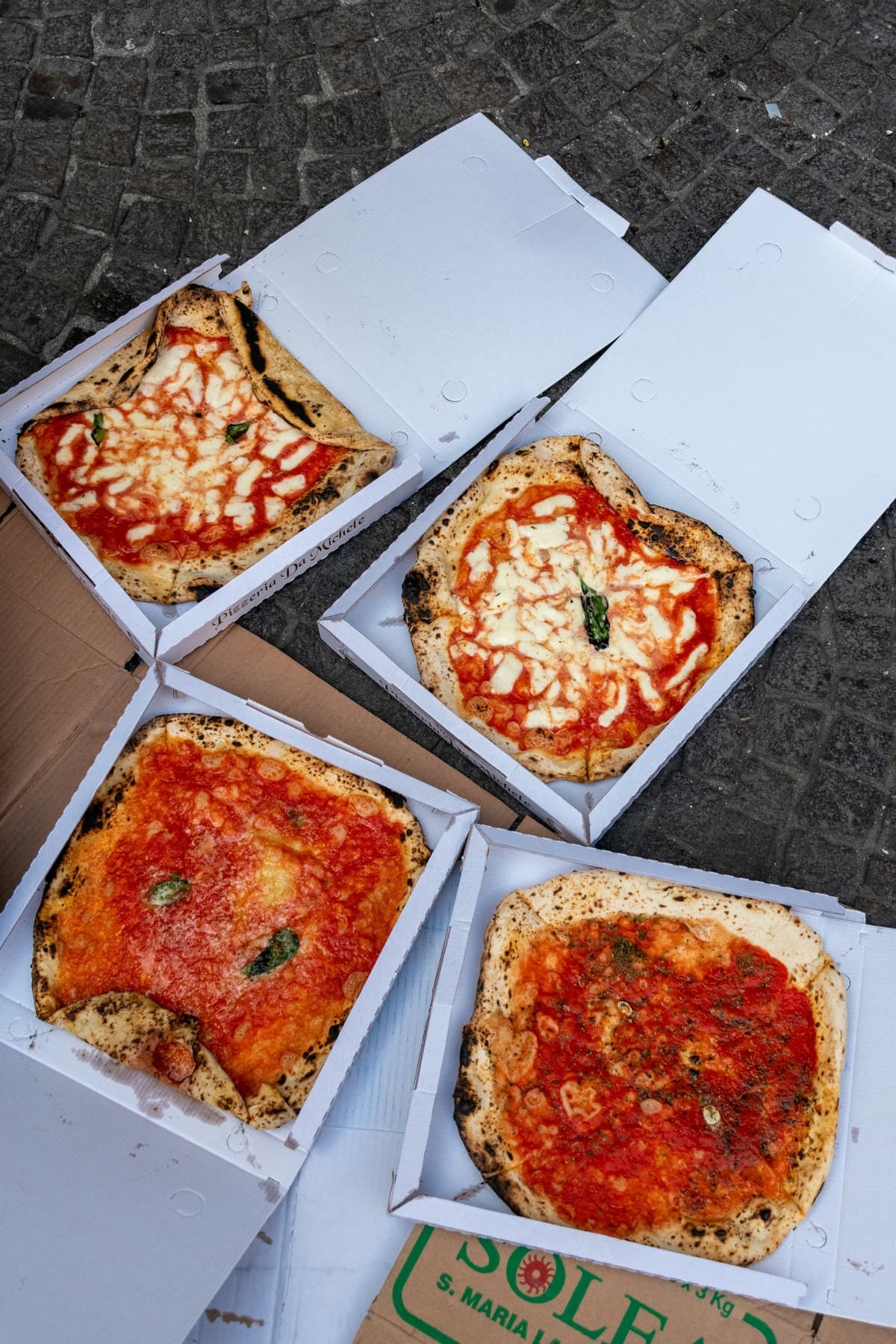Quatre pizzas dans leurs cartons, posées à même les pavés : deux Margherita, deux Marinara.