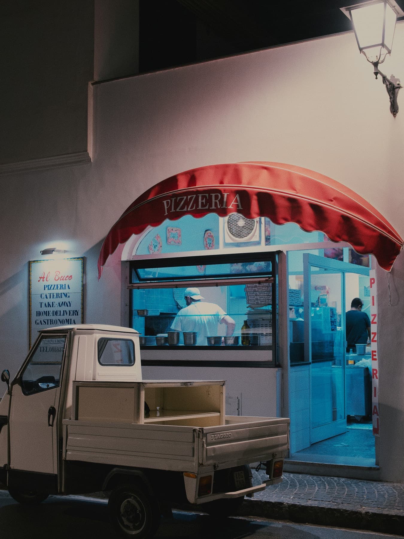 Pizzeria le soir, store rouge, vitrine allumée, un petit camion blanc le long du trottoir.