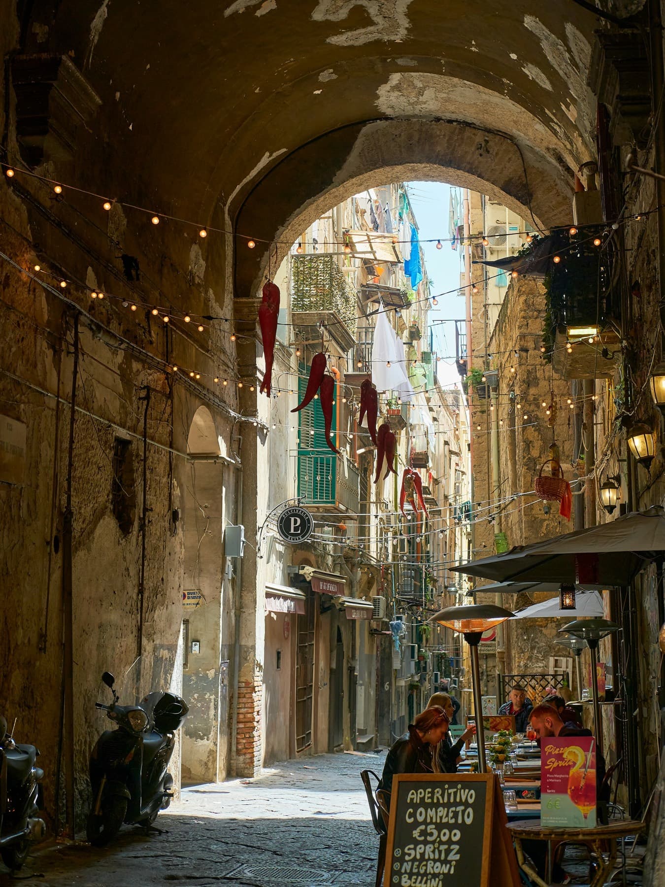Ruelle de Naples vue sous une voûte de pierre : linge aux balcons, lumières tendues d'un mur à l'autre.